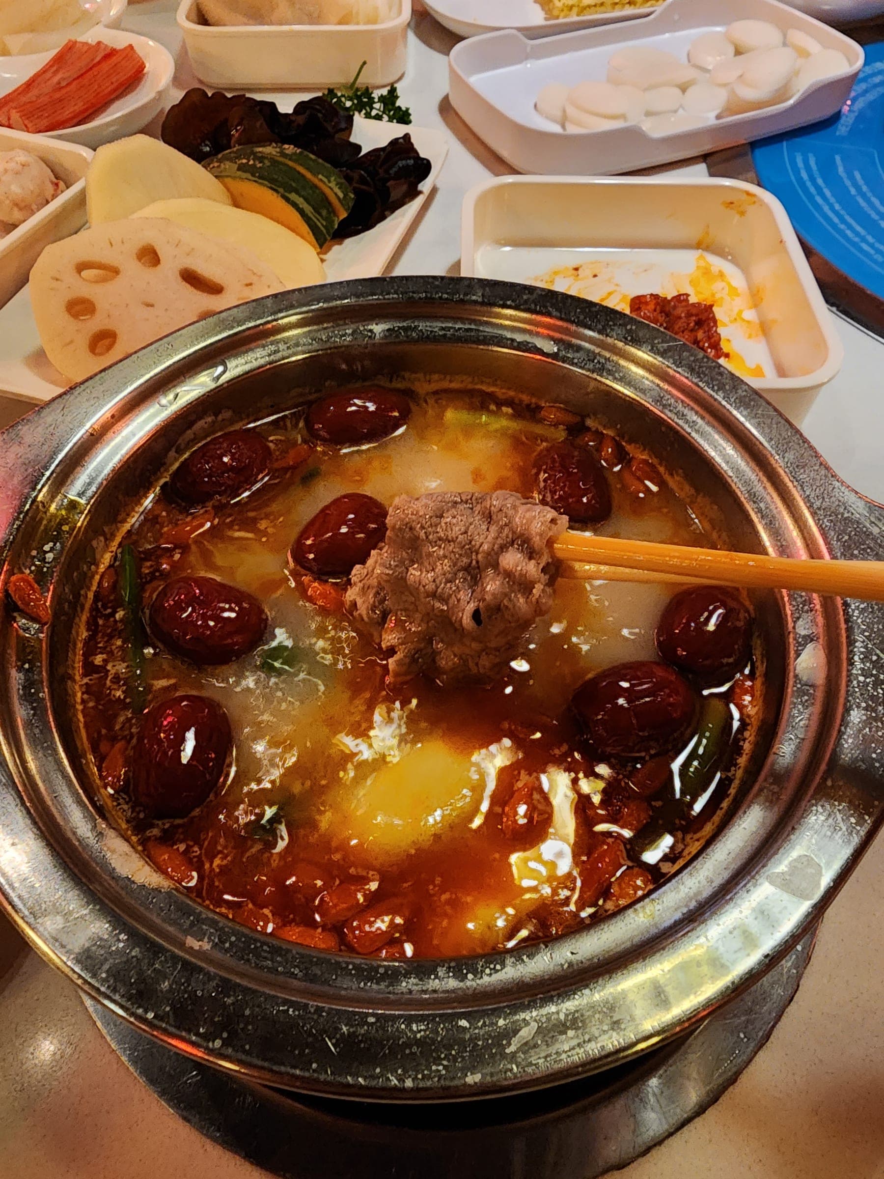 Chopsticks lifting a piece of beef out of a bubbling pot of chili broth studded with red dates and green chilies.