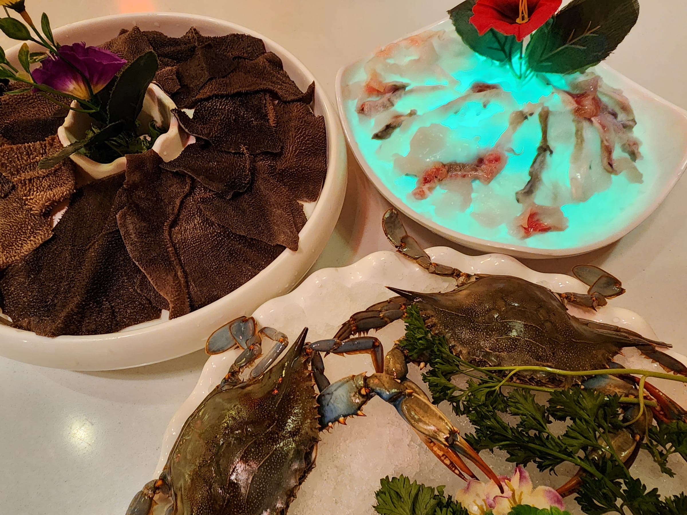 A bowl of sliced beef tripe and a lit platter of thin-sliced fish beside whole blue crabs resting on crushed ice.