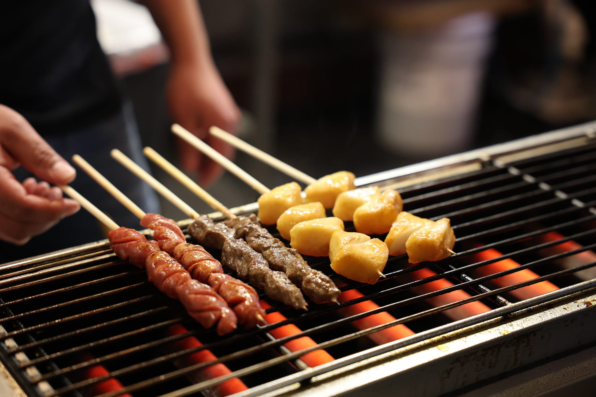 Skewers of sausage, lamb and rice cake cooking over a long charcoal grill.