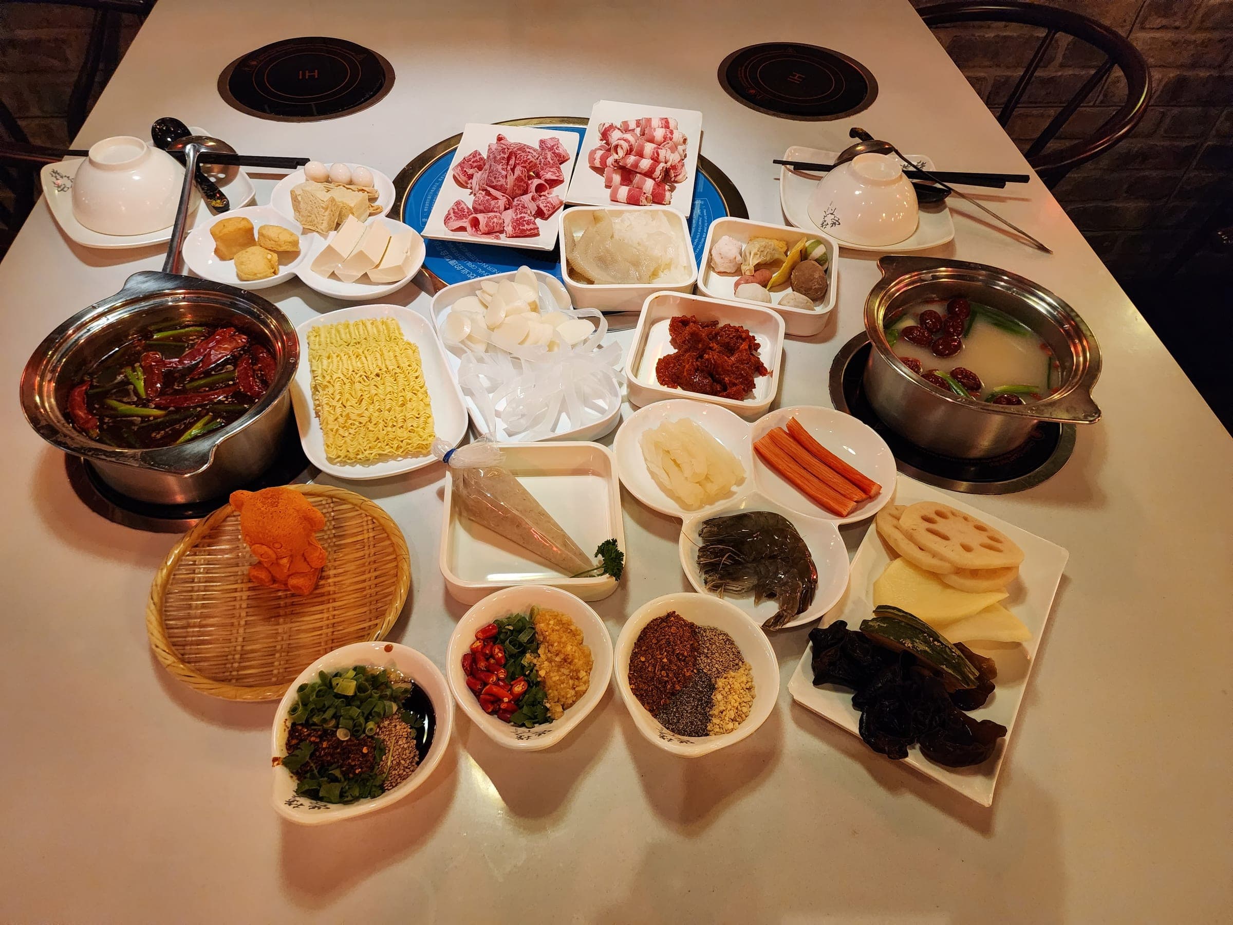 Overhead view of a spicy mala pot and a clear herbal pot surrounded by dishes of noodles, lotus root, mushrooms, carrots and dipping sauces.