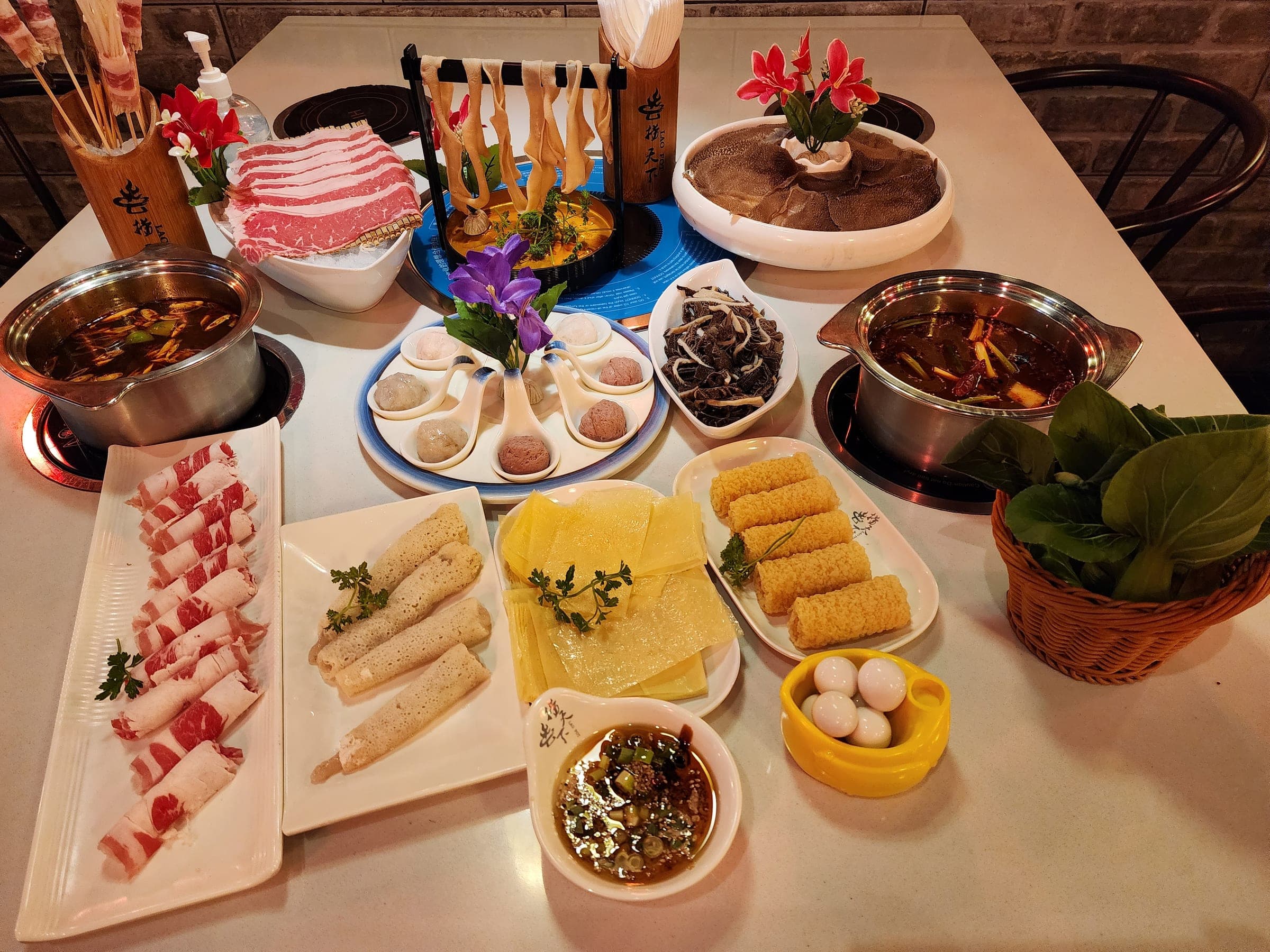 A table for four with two hot pots, rolled beef and pork, fish balls, tofu skin, quail eggs and greens laid out around them.