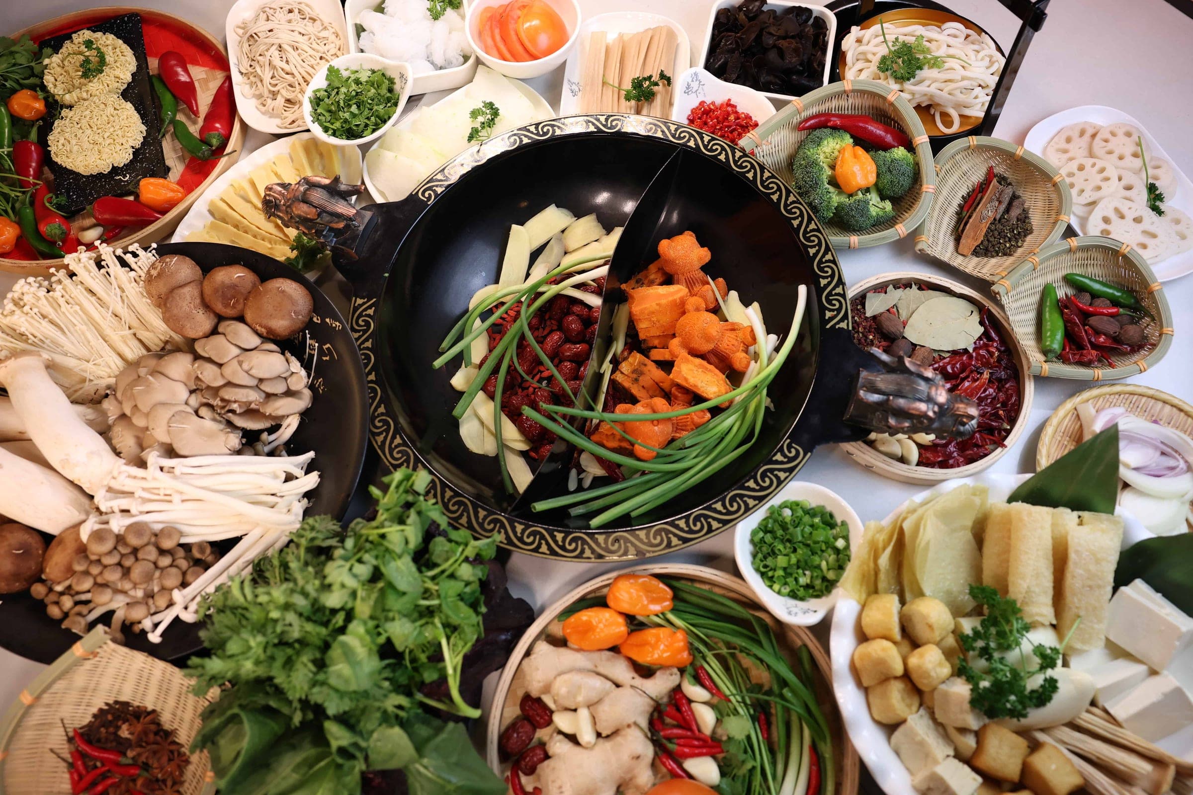 An overhead spread of hot pot ingredients around a black wok: mushrooms, greens, tofu, noodles, dried chilies and sliced meats.